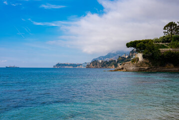 Fototapeta premium Turquoise Mediterranean meets rocky pine cliffs near Monaco, with Le Rocher and high rises on the mountainside, terraced gardens, clouds, and a lone ship on the horizon.
