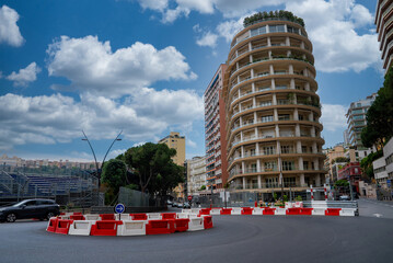 Street level view in Monaco shows a sweeping curve with barriers, a roundabout sign, a terraced cylindrical apartment tower, and grandstands under midday clouds. © Aerial Film Studio