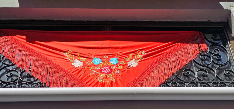 Red embroidered fringed shawl draped over ornate wrought iron balcony, showcasing traditional Andalusian textile decoration in bright sunlight