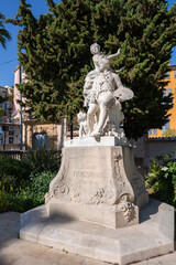 Fototapeta premium Stone statue of a painter with palette on ornate plinth in Grasse, France. Midday light casts crisp shadows on sculpted figures and floral reliefs in a small square.