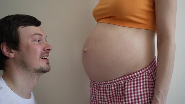 Caucasian man using a glass like a stethoscope to listen to his unborn baby's heartbeat inside his pregnant wife's womb, showing a range of comical emotions from curiosity to joy