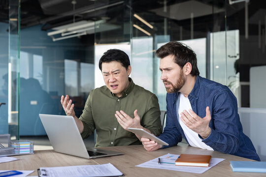 Two diverse businessmen feeling angry and confused while collaborating and facing a severe technical error or problem, on a laptop with documents on the modern office desk