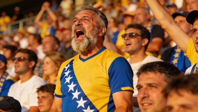 Shouting bearded senior fan in yellow-blue jersey with diagonal star stripe standing on bleachers
