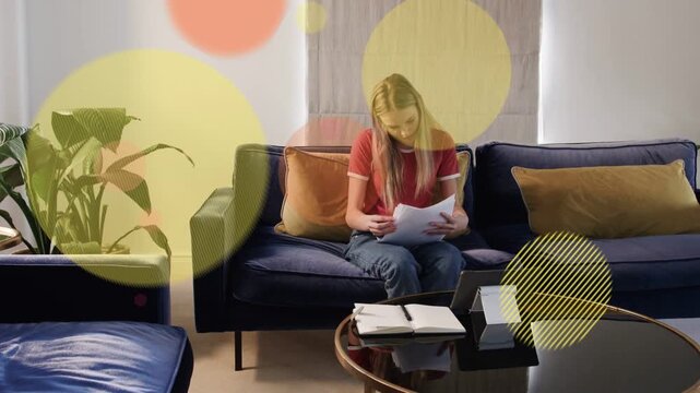 Vertical video: Picking papers, woman in red shirt and jeans reviewing on sofa at home with tablet