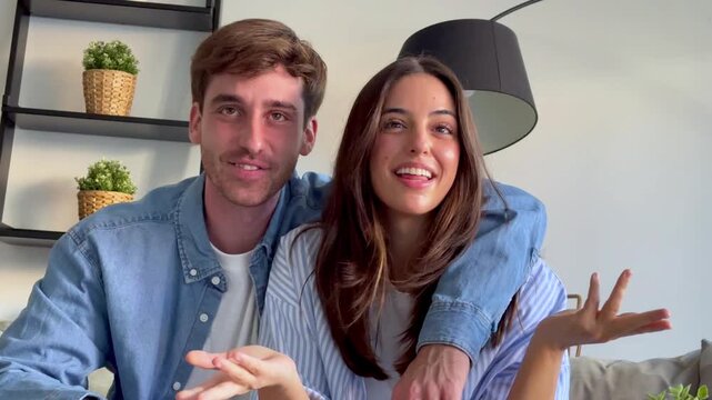 Joyful young caucasian couple sitting at home and happily talking on a video call with family, embracing each other while smiling, laughing, and gesturing in their modern living room