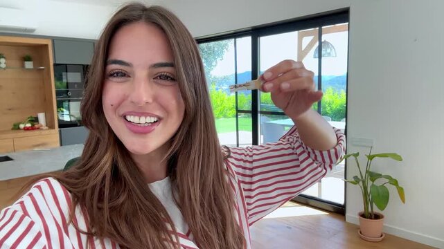 First-person view of a smiling young woman proudly displaying the keys to her new apartment, celebrating a fresh start and the achievement of homeownership in her modern living space