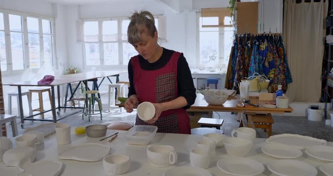 Artisan's Craft in Studio: An artisan, diligently focused, refines a piece of pristine pottery amidst a sunlit studio filled with various ceramic creations.