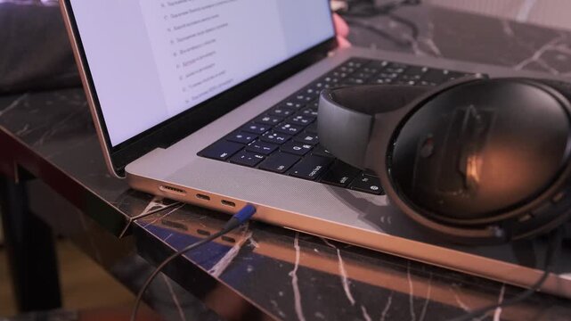 Close-up of hand plugging headphone jack into laptop audio port on desk, showing wired connection setup, sound device usage, everyday technology interaction, home workspace environment