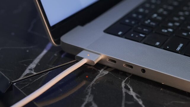 Close-up of hand attaching magnetic charging cable to laptop port on desk, demonstrating snap-in connector, modern charging technology, safe power connection, everyday device usage