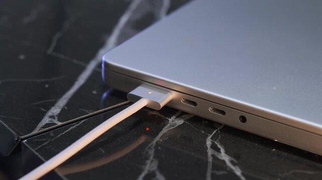 Close-up of hand attaching magnetic charging cable to closed laptop port on desk, showing snap-in connector, safe power connection, modern device charging, everyday technology usage