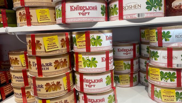 Assorted round boxes of the famous Roshen Kyiv Cake and Grillage Cake with their signature chestnut leaf design on shelves in a local grocery store in Kyiv.