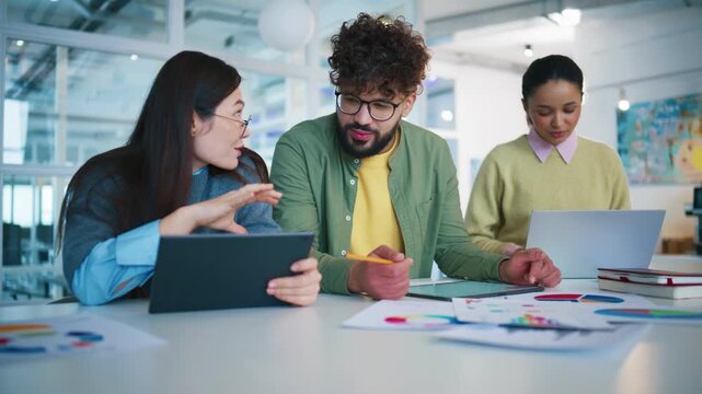 Creative planning session at shared desk. Asian woman presenting ideas on tablet device. Man listening with focus. Pretty woman standing besides team and typing on laptop in contemporary office.