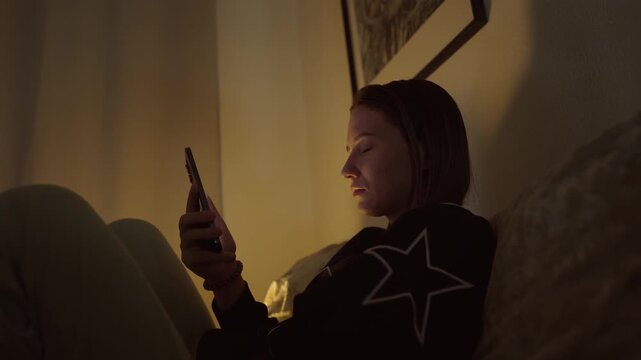Girl doom scrolling her smartphone on a coach at night. Social network and phone addiction concept. Cozy home ambient light