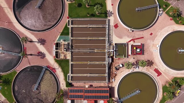 Overhead view of municipal wastewater treatment facility showing rectangular aeration basin with parallel lanes, four circular primary and secondary clarifiers with rotating sludge scrapers, solar pan