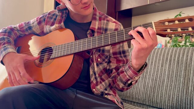 Young man in plaid shirt and glasses playing acoustic classical guitar on a grey couch, learning new chords at home