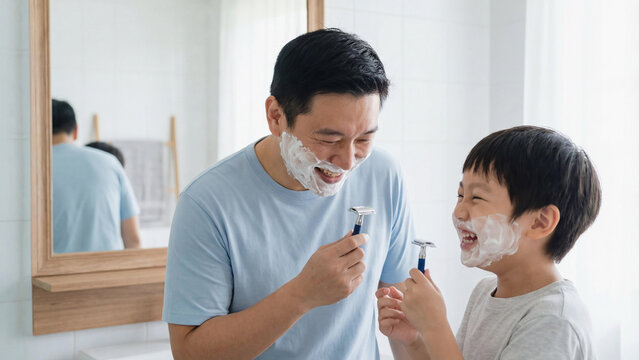 Happy Asian father and young son bonding laughing while pretend shaving in a bathroom. White foam on faces and holding razors. Family morning routine and joy. Fatherhood and parenting.