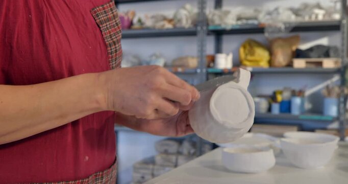 Crafting a moment: An artisan's careful touch shapes a plain white ceramic teapot and its delicate cups in a sunlit workshop, showcasing the beauty of pottery.