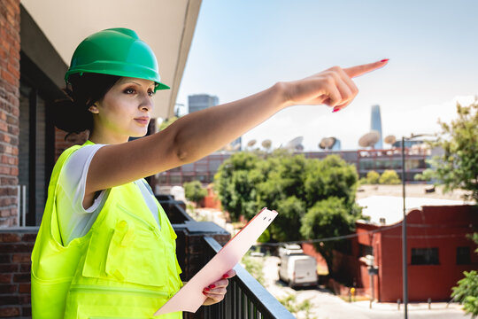 Young female safety inspector wearing green safety helmet and yellow safety vest, holding pink folder while supervising construction site safety and giving instructions from balcony