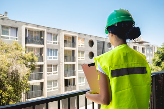 Young female safety inspector wearing green safety helmet and yellow safety vest, holding pink folder while supervising construction site safety and giving instructions from balcony