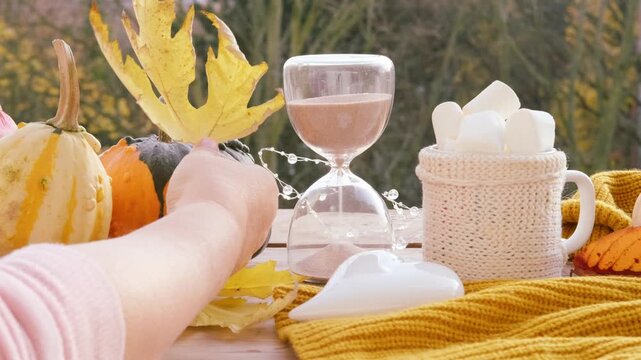 Autumn view with woman's hand holding yellow maple leaves, pumpkins on table in garden, sand in an hourglass, nature and changing seasons