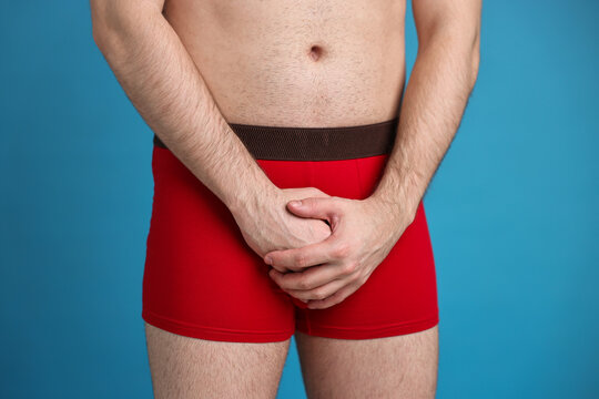 Urology. Man in red underwear covering his groin with hands on light blue background, closeup