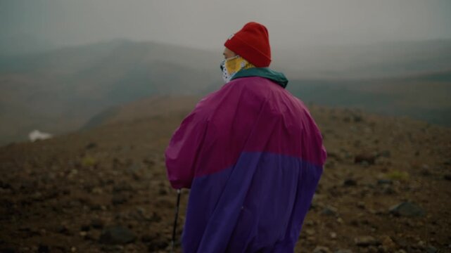 Young man in raincoat and in buff goes with trekking poles along rocky ridge high in mountains. Back view. Slow motion. Concept of privacy and contemplation