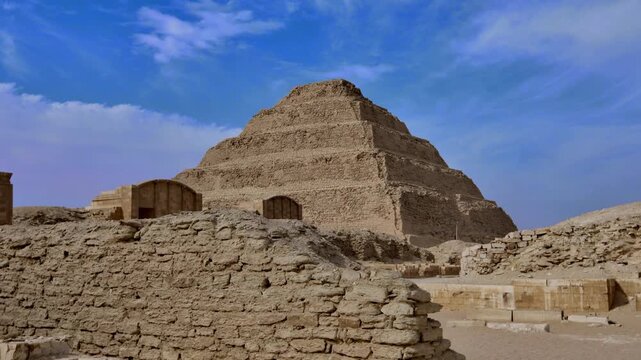 World's oldest pyramid, Step Pyramid of Djoser at Saqqara, Egypt, rising above ancient ruins and stone blocks with arched entrance wall under blue sky.