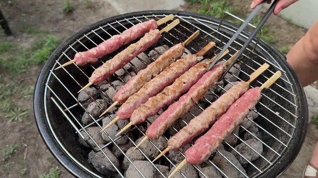 Outdoor charcoal grill with raw minced meat skewers, hand turning kebab sticks with metal tongs, casual backyard barbecue cooking, summer picnic meal prep