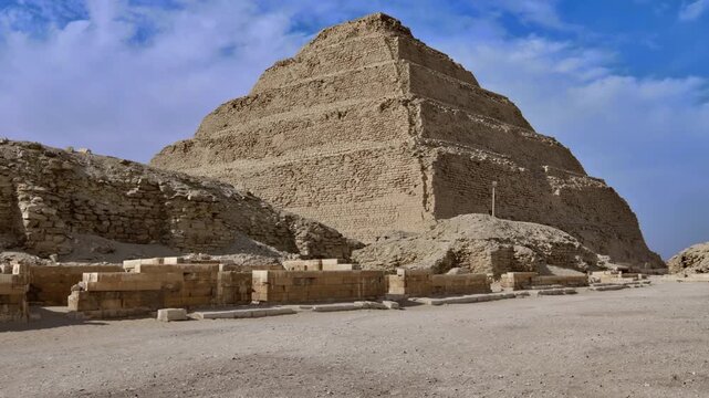 Massive six-stepped limestone pyramid of pharaoh Djoser at Saqqara necropolis, Egypt, with ancient enclosure wall, ruins and sandy desert landscape.