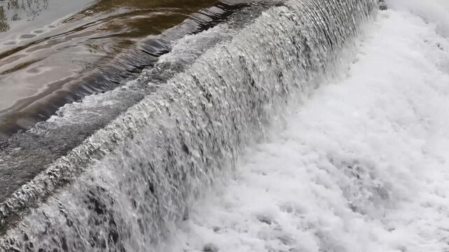 Rushing water flowing over a concrete weir, creating a turbulent cascade and white foam. Concept of clean energy and natural power