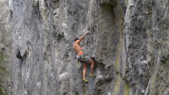 Shirtless young climber works through a steep limestone section beneath a small overhang, using body tension and careful footwork. Power, concentration and technical movement during a demanding climb.