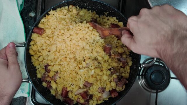 Top down view of hands cooking traditional spanish migas with salchicha and longaniza inside a black frying pan on a hot gas stove using a wooden spoon at home
