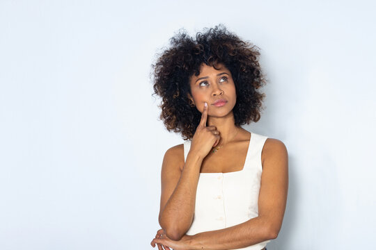 Young woman with natural curly hair posing thoughtfully, finger on cheek, reflective expression, light background, modern lifestyle style