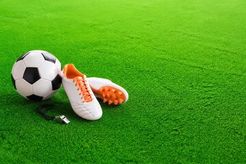 Naklejka premium Soccer ball and white cleats with orange accents placed on vibrant green artificial turf, whistle lying beside the shoes in a sports setting