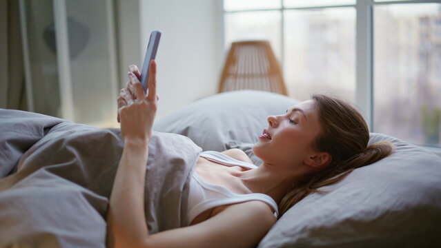 Awaking girl chatting smartphone in bed closeup. Dreamy brunette rejoicing sms