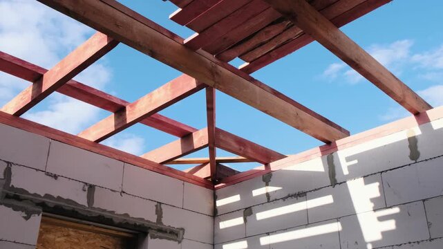 Low angle view of wooden roof frame on unfinished house with sunlight casting shadows on block walls, blue sky above, highlighting construction process, carpentry work and residential building
