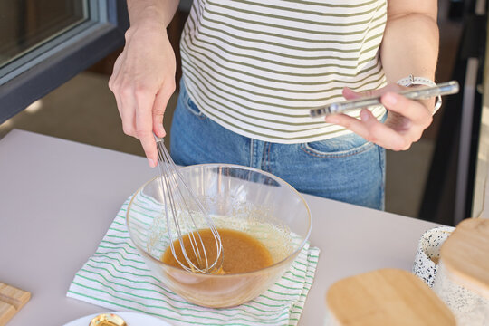 Woman mixes batter with whisk in glass bowl while holding smartphone