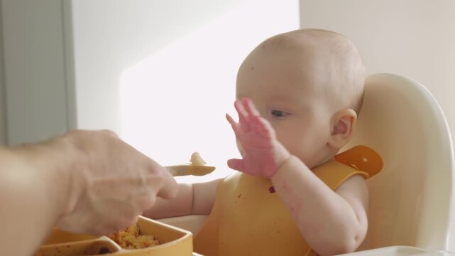Baby sits in a high chair and happily eating pureed food. Concept of early childhood and developmental milestones is conveyed. Child tasting vegetables at the table, discovering new food.