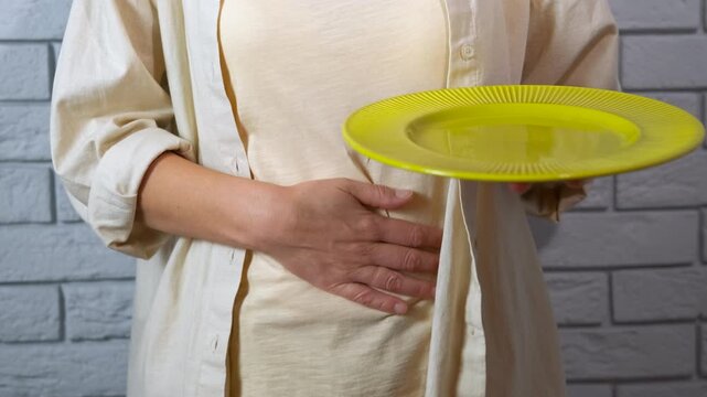 Woman holding an empty plate experiencing abdominal pain. Woman wearing casual clothes holding an empty yellow plate while massaging her abdomen due to stomach pain, possibly from hunger, indigestion