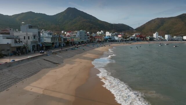 A coastal town of Nhon Hai, Quy Nhon, Binh Dinh, Vietnam with mountains in the background. The beach is lined with buildings and boats are visible in the water.