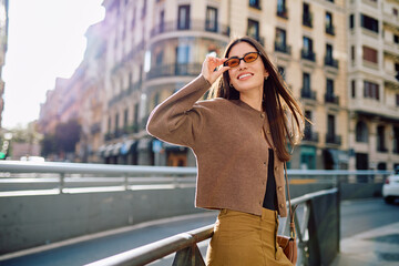 Fototapeta premium Young woman smiling, standing on a city street, lifting her sunglasses, enjoying the urban outdoor lifestyle and sunny day