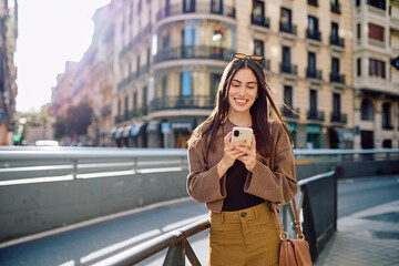Obraz premium Happy young woman smiling, checking phone messages while walking along a sunny city street, enjoying urban connectivity