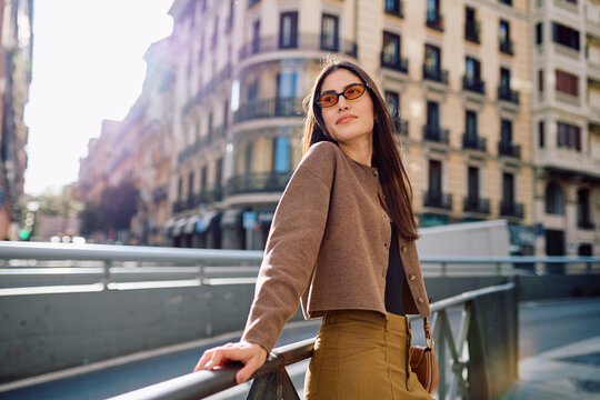 Young woman posing outdoors, leaning on a railing. She is enjoying urban lifestyle and modern street fashion