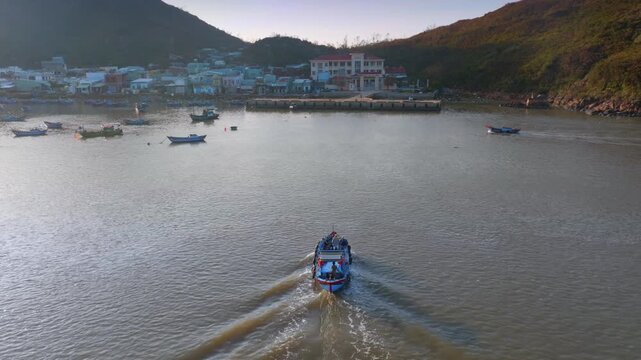 A fishing boat with several people on board travels through the water near a coastal town iof Hai Minh, Quy Nhon, Binh Dinh, Vietnam. The boat is leaving a wake as it moves.
