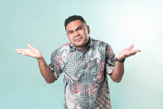 Portrait of a Southeast Asian man wearing batik shirt shrugging his shoulders with a confused and doubtful expression against a teal backdrop.
