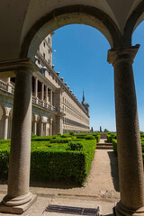 Obraz premium Framed view through stone arches of the manicured gardens and classical architecture of the Royal Monastery of San Lorenzo de El Escorial, a UNESCO World Heritage site in Madrid, Spain.