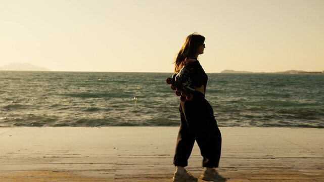 Full length young woman in black carrying roller skates along seafront at warm golden sunset. Turquoise waves, coastal city on horizon. Active outdoor summer lifestyle and freedom.

