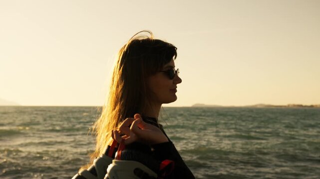 Portrait of young woman with wind-blown hair holding roller skates at golden sunset by the sea. Warm backlight, islands on horizon. Confident stylish active summer lifestyle.
