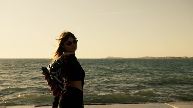 Young woman in black carries roller skates on shoulder along seafront at golden sunset. Wind in hair, warm sea light, coastal city on horizon. Active outdoor summer lifestyle.
