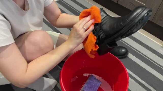 Caucasian woman drying and polishing black boot with orange cloth over red bucket, smoothing leather, creating shine, finishing touch sequence, tidy apartment scene, satisfying cleaning closeups